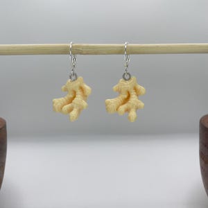 Op de afbeelding: Een paar crèmekleurige oorbellen in de vorm van koraal met zilverkleurige haken. De oorbellen hangen aan een houten deuvel tegen een witte achtergrond. De oorbellen hebben een textuur, organisch uiterlijk.