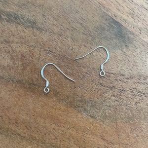 May include: Two silver earring hooks, a type of jewelry finding, are shown on a wooden surface.