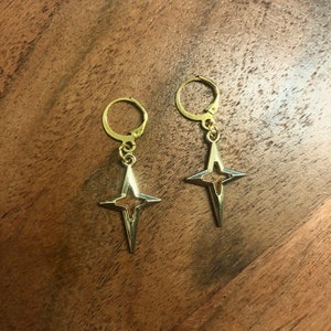 May include: A pair of gold hoop earrings with a dangling star charm. The star is open in the center and has a pointed design.