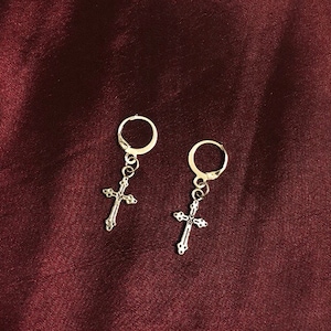 May include: A pair of silver hoop earrings with a dangling cross charm. The earrings are on a dark red background.