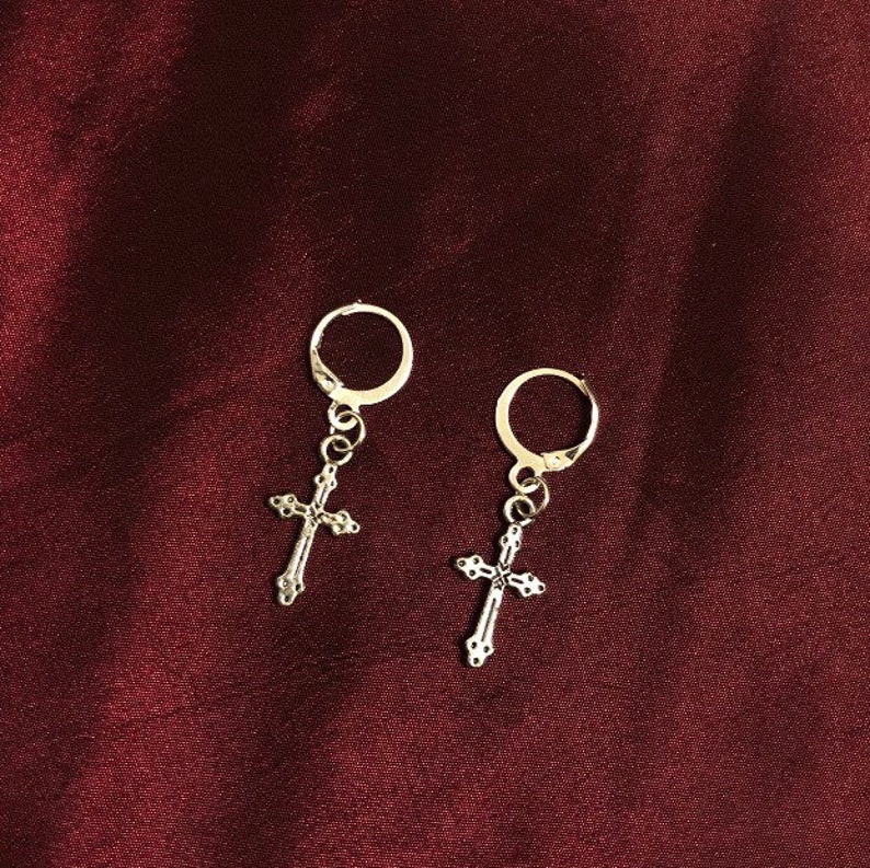 May include: A pair of silver hoop earrings with a dangling cross charm. The earrings are on a dark red background.