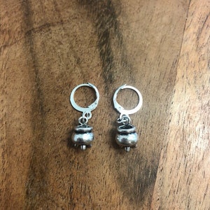 May include: A pair of silver hoop earrings with a small silver cauldron charm hanging from each hoop.