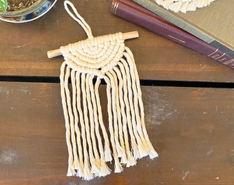 May include: A small, cream-colored macrame wall hanging with a wooden dowel at the top and fringe at the bottom.