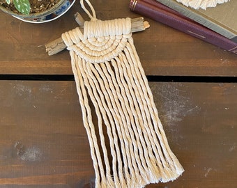 May include: A small macrame wall hanging with a natural wood branch and a cream colored fringe. The hanging is in the shape of a half circle.