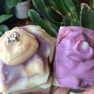 May include: Two bars of handmade soap, one with a swirl of purple and white and a small silver goat charm on top, and the other with a swirl of purple and white.