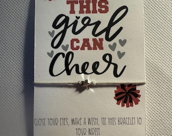Peut inclure: Une carte blanche avec le texte "THIS girl CAN Cheer" en lettres noires et rouges, avec de petits coeurs gris et des graphiques de pompons. Un bracelet avec un charme en forme d'étoile est attaché. La carte comprend des instructions.