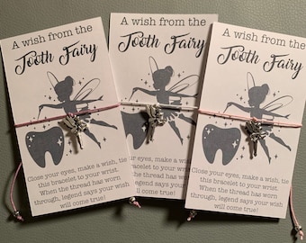 May include: Three white cards with pink string attached to each. The cards feature a tooth fairy illustration and the text "A wish from the Tooth Fairy". Each card has a tooth charm and a message that reads "Close your eyes, make a wish, tie this bracelet to your wrist. When the thread has worn through, legend says your wish will come true!"