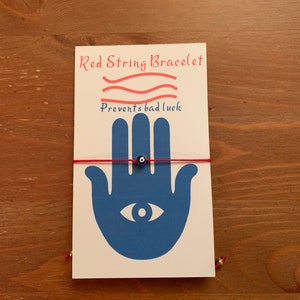 Op de afbeelding: Rood draad armband met een blauwe kwade oog bedel. De kaart zegt "Rood draad armband" en "Voorkomt ongeluk".