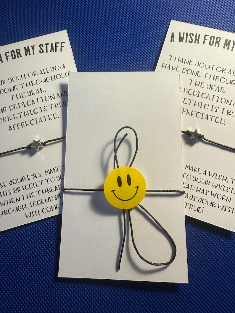 May include: Two white cards with black text and a black string with a yellow smiley face charm and a silver star charm. The text on the cards reads "Thank you for all you have done throughout the year. Your dedication and work ethic is truly appreciated." and "Make a wish, tie to your wrist. The thread has worn, says your wish true!"
