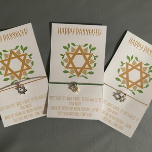 May include: Three white cards with the text "Happy Passover" printed on them. Each card has a gold Star of David with green leaves around it. Each card has a string bracelet attached with a silver Star of David charm. The cards have instructions to make a wish and tie the bracelet on your wrist.