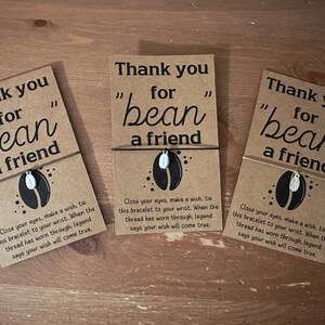 May include: Three brown cards with the text "Thank you for "bean" a friend" printed on them. Each card has a small silver charm of a coffee bean attached to a brown string.