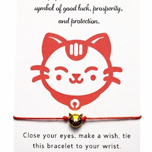 May include: Red Maneki-neko cat charm bracelet. The card reads: "The Lucky cat, known as Maneki-neko in Japanese, is a traditional symbol of good luck, prosperity, and protection." The bracelet is tied with a red string.