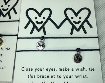 Può includere: Due braccialetti dell'amicizia con filo nero e ciondoli argentati "BFF" sono esposti su biglietti con la scritta "Besties". Le carte presentano grafiche di cuori sorridenti e testo che descrive lo scopo del braccialetto.