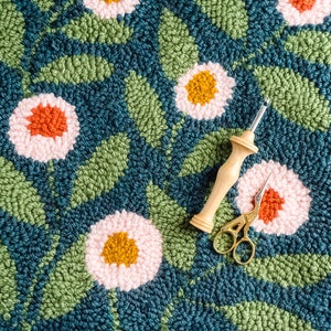 May include: A close-up of a rug with a floral design. The rug is made of yarn and has a variety of colors, including pink, green, and blue. A wooden needle and a pair of gold scissors are on top of the rug.