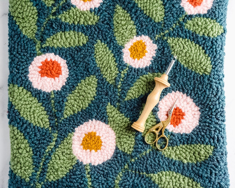May include: A close-up of a rug with a floral design. The rug is made of yarn and has a variety of colors, including pink, green, and blue. A wooden needle and a pair of gold scissors are on top of the rug.