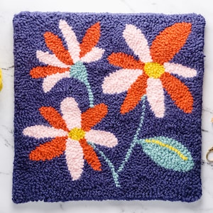 May include: A close-up of a needlepoint rug featuring a floral design. The rug depicts three large flowers with pink, orange, and yellow petals. The background of the rug is a deep blue colour.