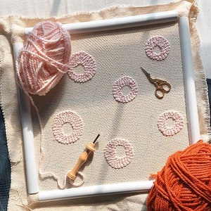 May include: A close-up of a pink yarn wrapped around a wooden embroidery hoop. The hoop is filled with a beige fabric with pink yarn circles stitched into it. A wooden needle and a pair of gold scissors are also visible. A ball of orange yarn is in the bottom right corner.