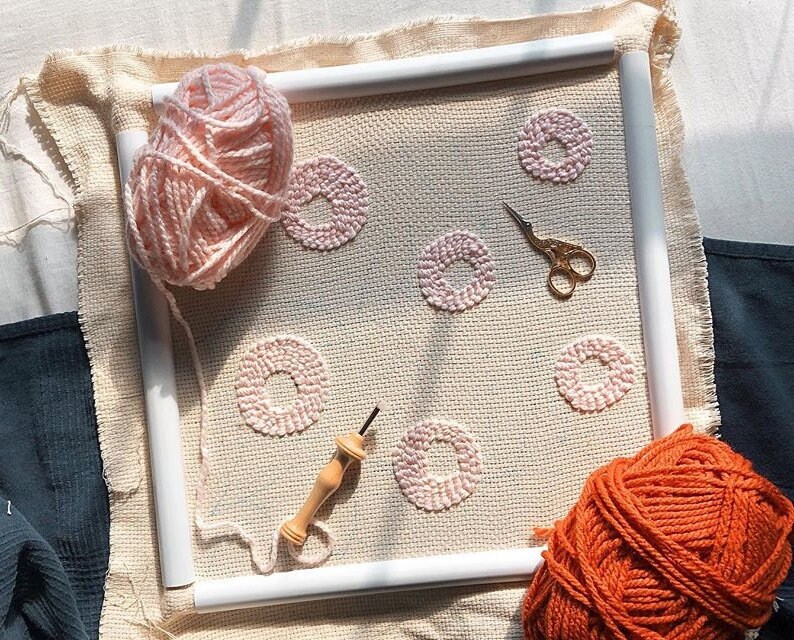 May include: A close-up of a pink yarn wrapped around a wooden embroidery hoop. The hoop is filled with a beige fabric with pink yarn circles stitched into it. A wooden needle and a pair of gold scissors are also visible. A ball of orange yarn is in the bottom right corner.