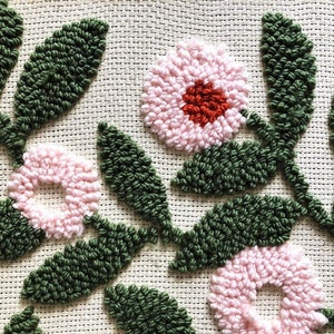 May include: Close-up of a hand-stitched embroidery featuring pink and green flowers with a red center. The flowers are made with a technique called punch needle embroidery.