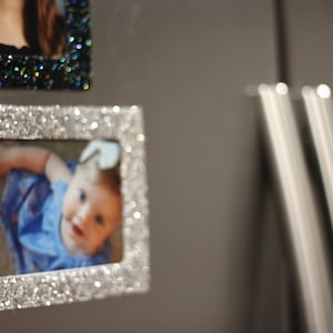 May include: A silver glitter picture frame with a photo of a baby wearing a blue and white outfit.
