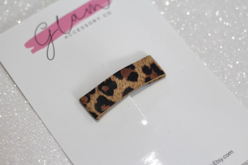 Puede incluir: Un peque&ntilde;o clip para el pelo con estampado de leopardo. El clip es marr&oacute;n y negro con un brillo sutil. El clip est&aacute; sobre un fondo blanco con el texto "Glam Accessory Co" escrito en rosa.