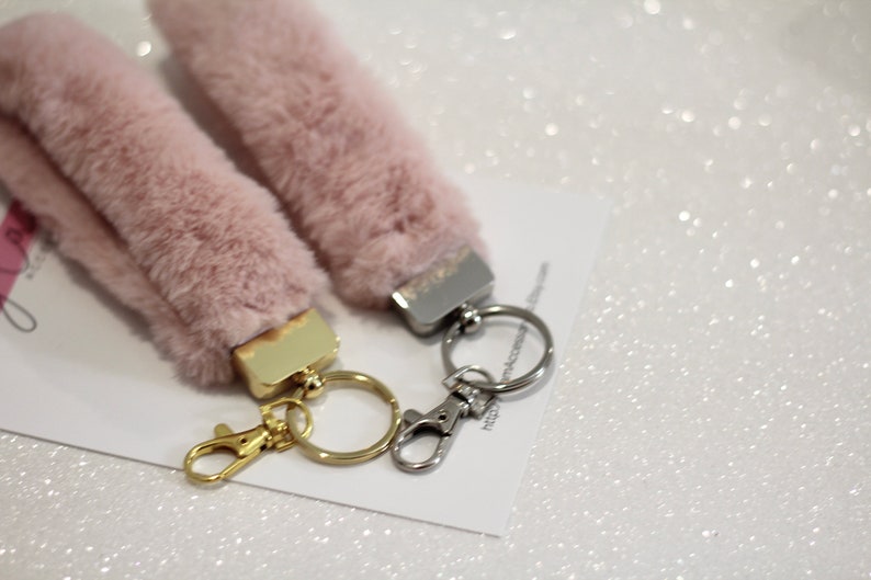 May include: Two fluffy pink keychains with gold and silver key rings. The keychains are made of faux fur and have a small metal charm attached to the top.