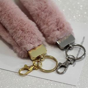May include: Two pink fluffy keychains with gold and silver key rings. The keychains are attached to a white card with the text "Glam4Accessor" and a website address.