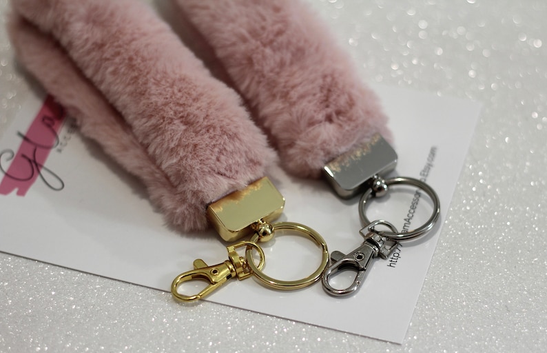 May include: Two pink fluffy keychains with gold and silver key rings. The keychains are attached to a white card with the text "Glam4Accessor" and a website address.