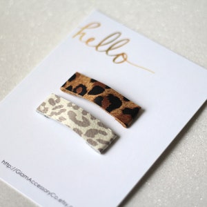 Puede incluir: Dos pinzas para el pelo de estampado de leopardo, una marr&oacute;n y una blanca, sobre una tarjeta blanca con el texto "hello" y una direcci&oacute;n de sitio web.