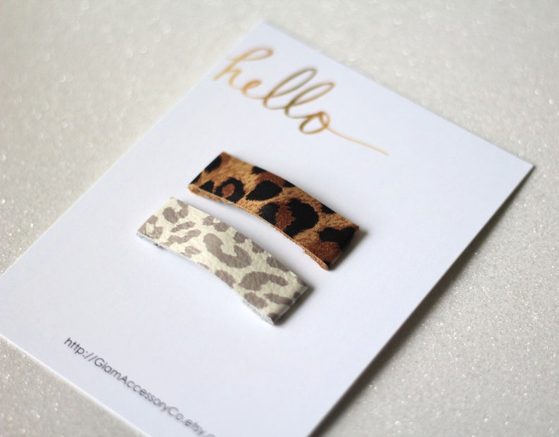 Puede incluir: Dos pinzas para el pelo de estampado de leopardo, una marr&oacute;n y una blanca, sobre una tarjeta blanca con el texto "hello" y una direcci&oacute;n de sitio web.