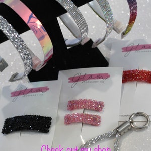 Puede incluir: Surtido de accesorios para el cabello brillantes, que incluyen diademas, pinzas y un llavero. Las diademas son plateadas, iridiscentes y de colores arco&iacute;ris. Las pinzas son negras y rosas. El llavero es plateado con un cord&oacute;n brillante. Los productos se muestran sobre un fondo blanco con el texto "Glam Accessory Co."