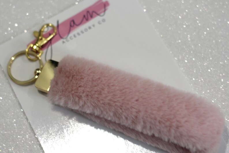 May include: A pink fluffy keychain with a gold key ring and a gold metal connector. The keychain has the text "Glam Accessory Co" on a white background.