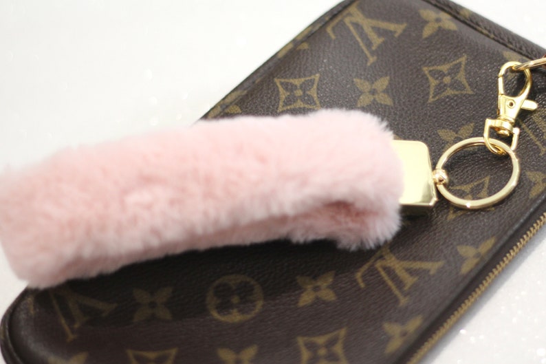 May include: A pink fluffy keychain charm attached to a gold key ring and a brown leather purse with a monogram pattern.
