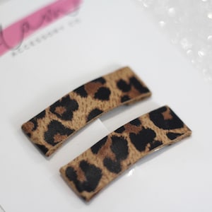 Puede incluir: Dos pinzas para el pelo de color marr&oacute;n y negro con estampado de leopardo. Las pinzas son rectangulares y tienen una forma ligeramente curvada.