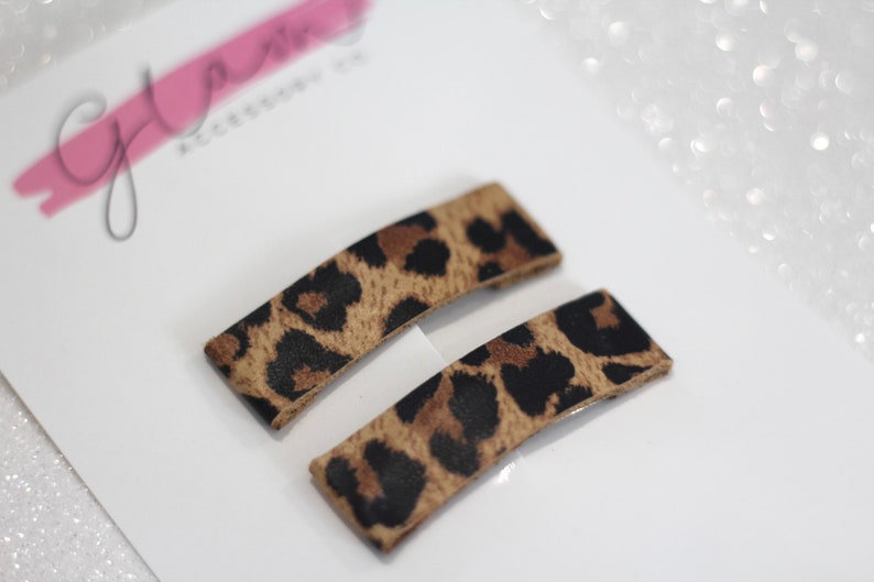 Puede incluir: Dos pinzas para el pelo de color marr&oacute;n y negro con estampado de leopardo. Las pinzas son rectangulares y tienen una forma ligeramente curvada.