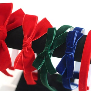 May include: Five velvet headbands with bows in different colors. The headbands are red, green, blue, and two are red. The headbands are displayed on a black stand.