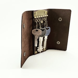 May include: A brown leather key holder with a brass clasp and several keys attached. The key holder is open, revealing the keys. The keys have different shapes and colors. The key holder is closed with two snap closures.