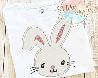 May include: A white t-shirt with a pink and gray embroidered bunny face. The bunny has pink ears, black eyes, and a pink nose.