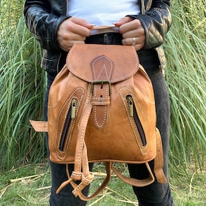 Puede incluir: Una mochila de cuero marrón con cierre de hebilla y dos bolsillos con cremallera. La mochila tiene costuras blancas y un cierre de cordón.