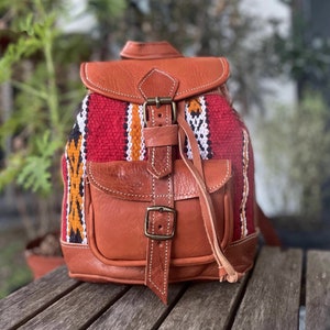May include: A small, brown leather backpack with a red, white and black woven pattern. The backpack has a flap with a buckle closure and a small front pocket.