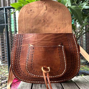 May include: A brown leather saddlebag with a flap closure and a front pocket. The bag features decorative stitching and a long adjustable strap. The interior is visible, showing the natural leather color. The bag is designed with a rustic, bohemian style.