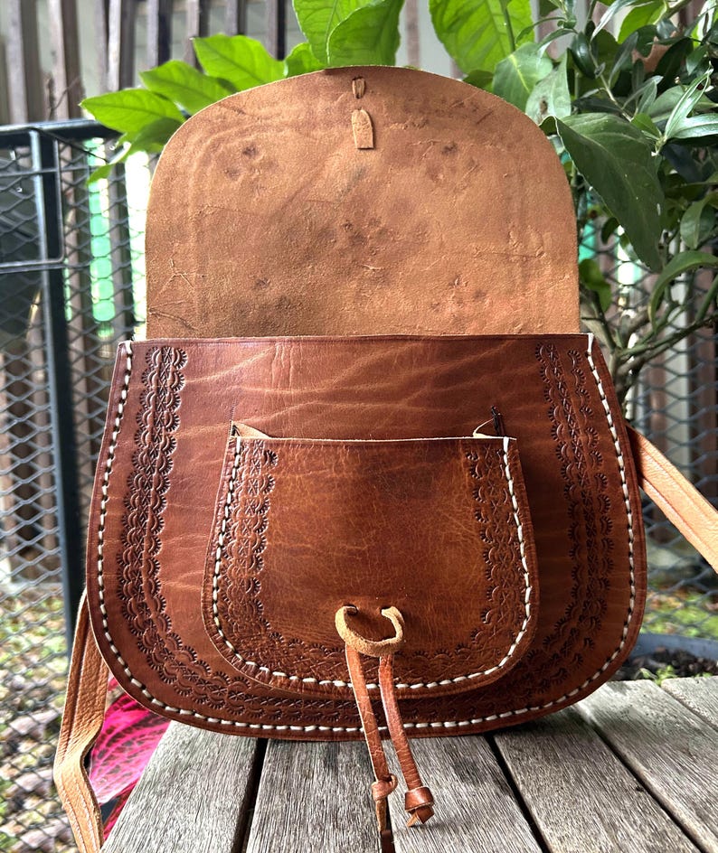 May include: A brown leather saddlebag with a flap closure and a front pocket. The bag features decorative stitching and a long adjustable strap. The interior is visible, showing the natural leather color. The bag is designed with a rustic, bohemian style.