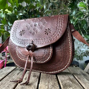 May include: A brown leather crossbody bag with floral and geometric embossed designs. The bag features a flap closure with a decorative toggle and a long adjustable strap. White stitching accents the edges of the bag.