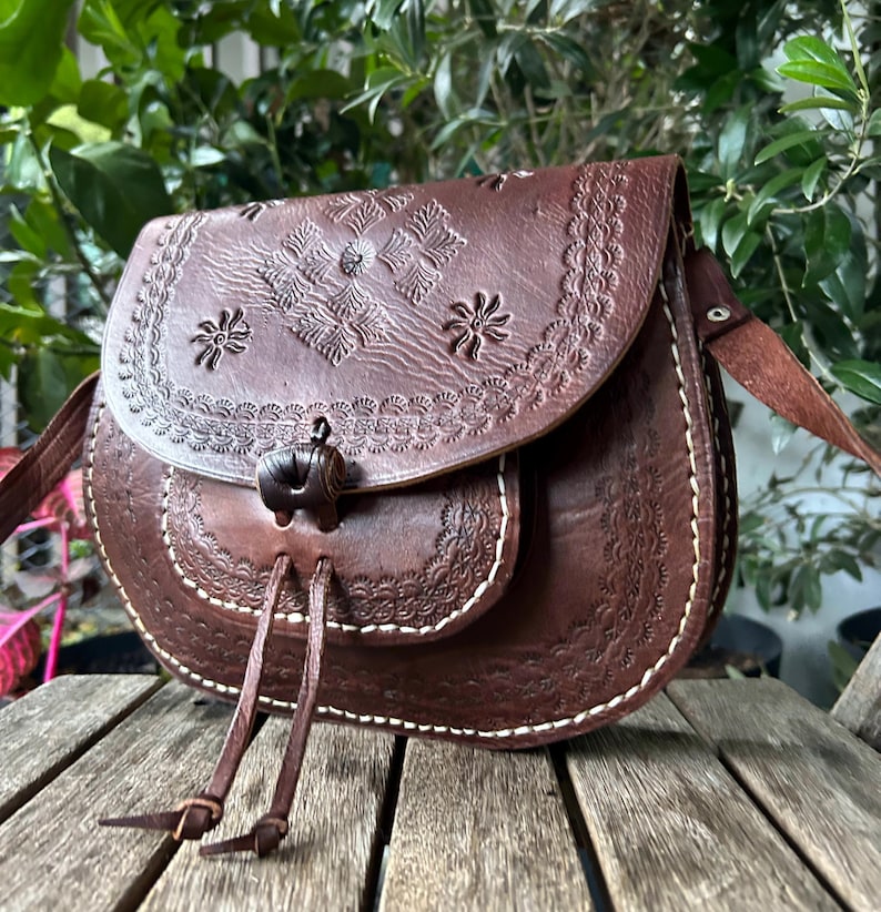 May include: A brown leather crossbody bag with floral and geometric embossed designs. The bag features a flap closure with a decorative toggle and a long adjustable strap. White stitching accents the edges of the bag.
