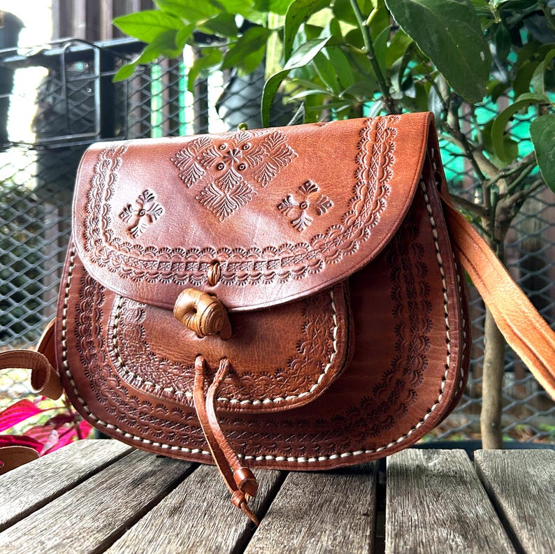 May include: A brown leather crossbody bag with intricate embossed floral patterns. The bag features a flap closure with a wooden toggle and a long, adjustable strap. The bag is approximately 10 inches wide and 8 inches tall.