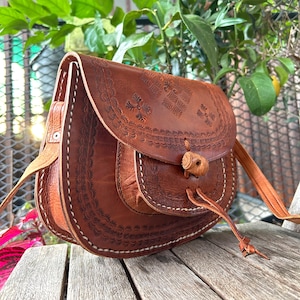 May include: A brown leather crossbody bag with intricate embossed floral patterns. The bag features a flap closure with a wooden toggle and a long, adjustable strap. White stitching accents the edges, and the bag appears to be handmade.