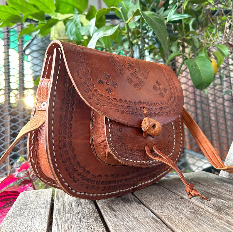 May include: A brown leather crossbody bag with intricate embossed floral patterns. The bag features a flap closure with a wooden toggle and a long, adjustable strap. White stitching accents the edges, and the bag appears to be handmade.