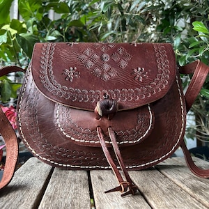 May include: A brown leather crossbody bag with a flap closure and a decorative floral pattern. The bag features a long adjustable strap and a drawstring closure. The bag is made of leather and has white stitching.