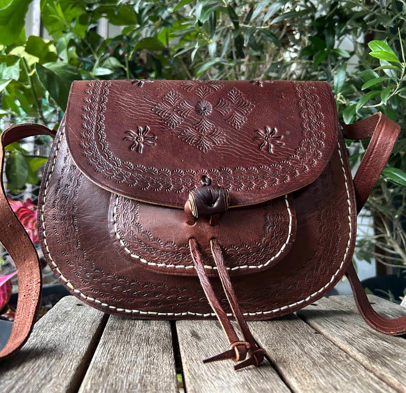 May include: A brown leather crossbody bag with a flap closure and a decorative floral pattern. The bag features a long adjustable strap and a drawstring closure. The bag is made of leather and has white stitching.