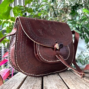 May include: A brown leather saddlebag with floral embossed detailing and white stitching. The bag features a flap closure with a wooden toggle and a long adjustable strap. The bag is sitting on a wooden surface.